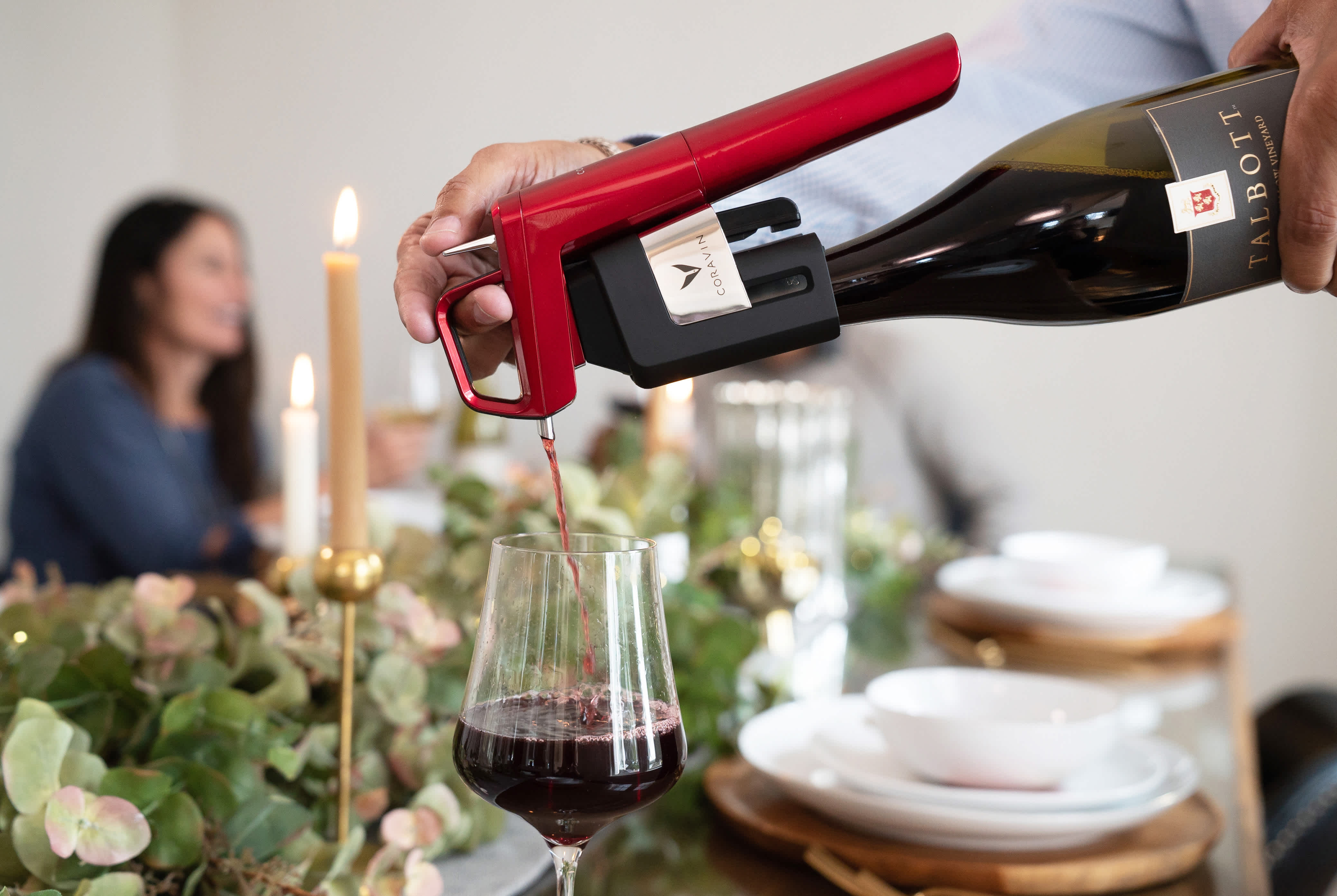 Lifestyle shot of wine being poured into a glass using a Coravin Model Six Wine Preservation System in Candy Apple Red. The wine glass is set on a table with candles, greenery, plates, and a woman in the background.