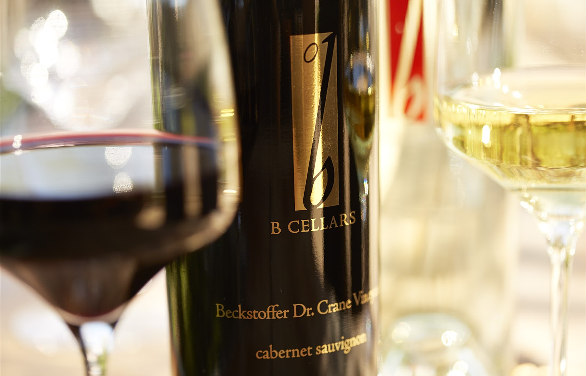 Close up shot of wine glasses with red wine and white wine, featuring a bottle of B Cellar wine.