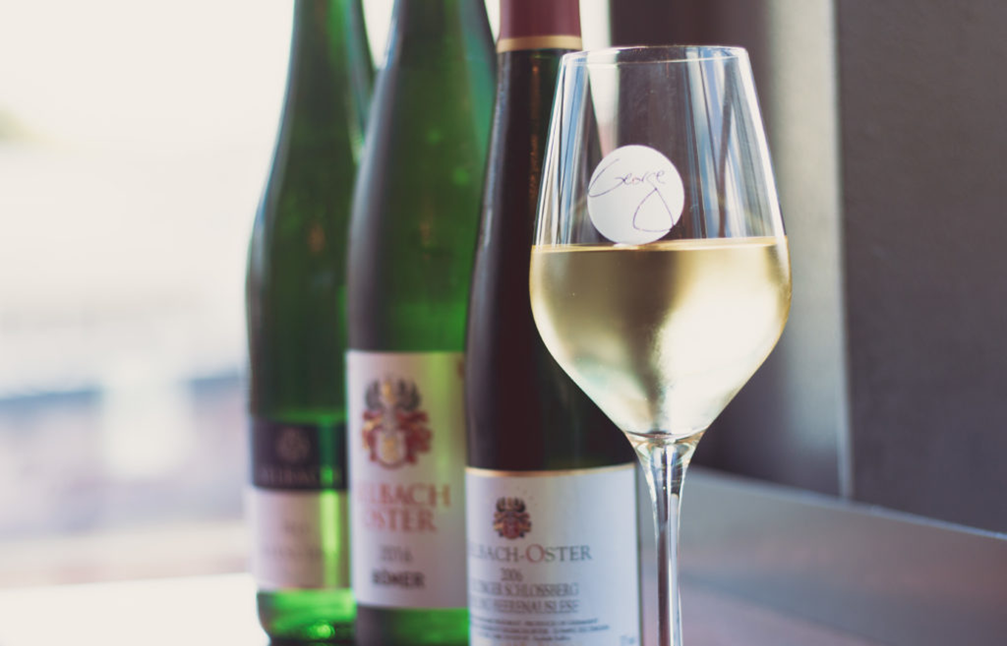 Een wijnglas van George Miliotes, gevuld met witte wijn, met wijnflessen op de achtergrond.
