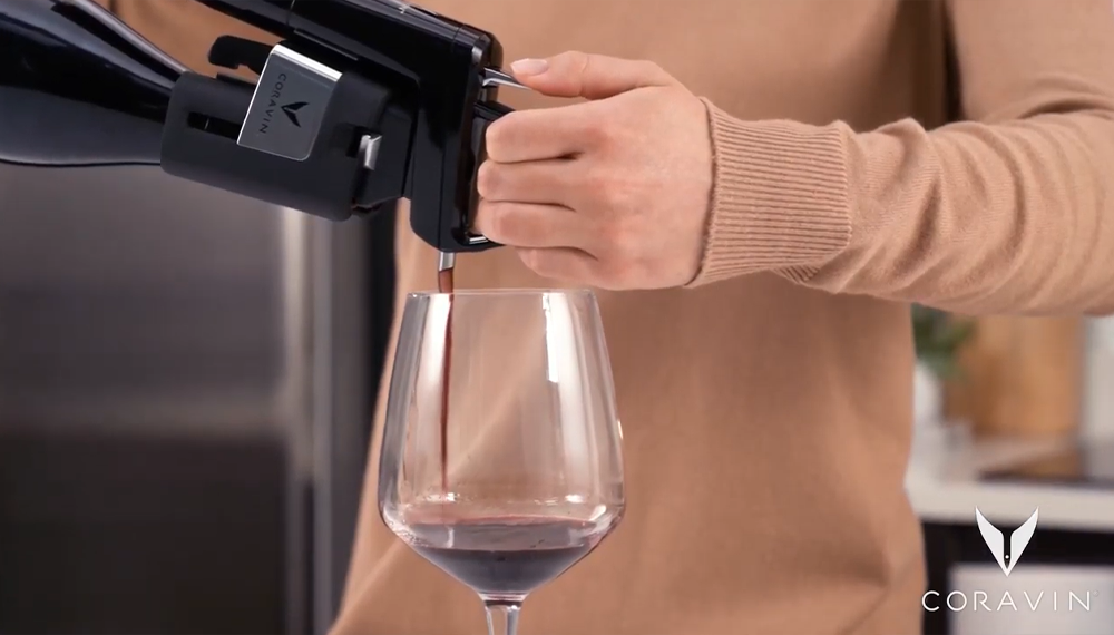 Primo piano di una donna che versa vino rosso in un bicchiere mediante un sistema di conservazione del vino Coravin.
