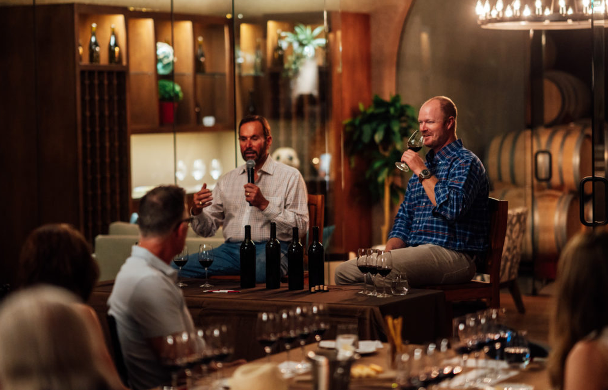 Two men talking in front of a crowd at a wine tasting in a wine cellar.