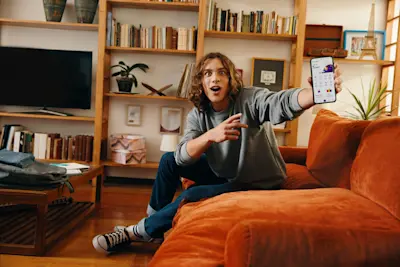 Joven sorprendido en el sillón señala la app de su cuenta en el celular