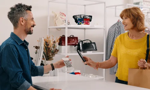Mujer paga con su teléfono celular en una tienda, mientras un hombre sostiene el código QR del comercio. 