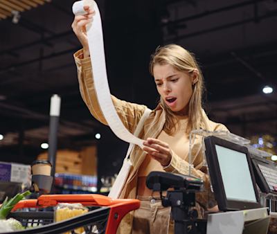 ¿Cómo varía el precio de un mismo producto según el tipo de comercio? Una mujer sostiene un ticket en la caja del supermercado y se muestra sorprendida por el monto que gastó.