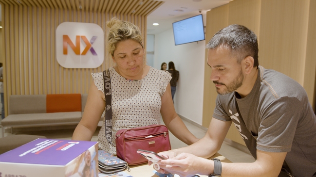 Nxer atiende a cliente en sucursal de Naranja X. Le muestra el teléfono; parece revisar la app Naranja X. 