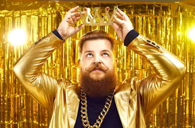 Hombre barbudo con saco dorado se pone una corona sobre fondo de cortina dorada