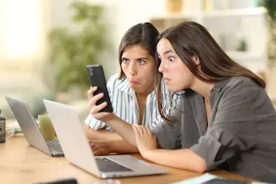 Dos mujeres sorprendidas leen un mensaje en el celular frente a la notebook