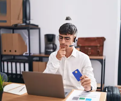 Hombre con auriculares duda frente a la notebook con una tarjeta en la mano
