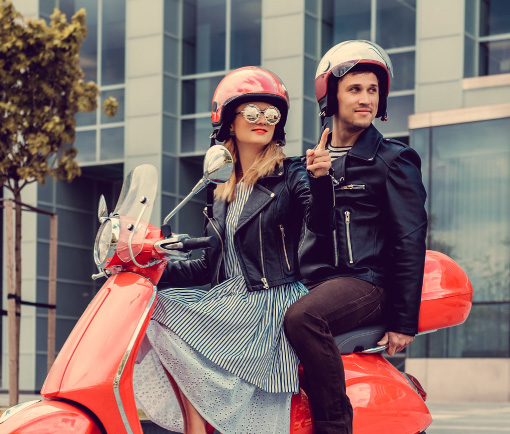 Hombre y mujer jóvenes con casco sobre una moto tipo Vespa color roja
