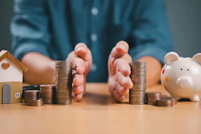 Manos que separan pilas de monedas entre una casa de madera y una alcancía