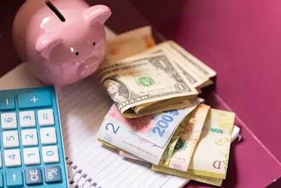 Alcancía rosa, calculadora y billetes de pesos y dólares sobre un cuaderno