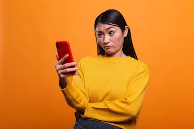 Mujer desconfiada mira de reojo su celular rojo sobre fondo naranja
