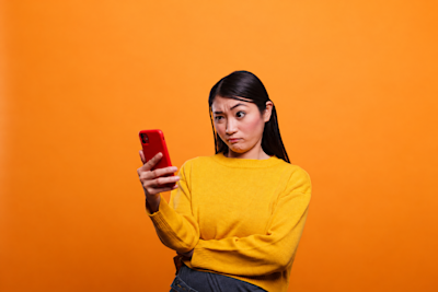 Mujer desconfiada mira de reojo su celular rojo sobre fondo naranja