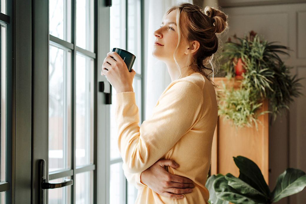 Eine junge Frau steht mit einem Kaffeebecher am Fenster in ihrer Wohnung und schaut hinaus
