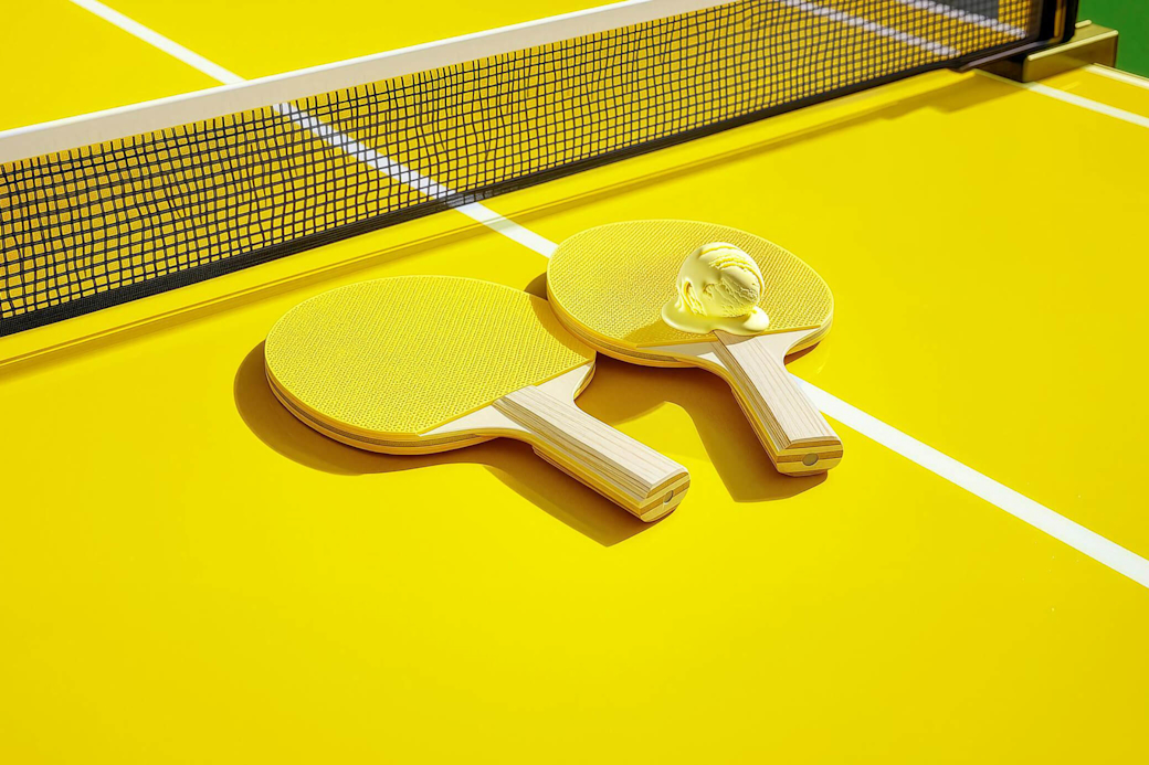 Zwei Tischtennisschläger liegen auf gelber Tischtennisplatte mit einer Eiskugel die an einen Tennisball erinnert - KI generiert