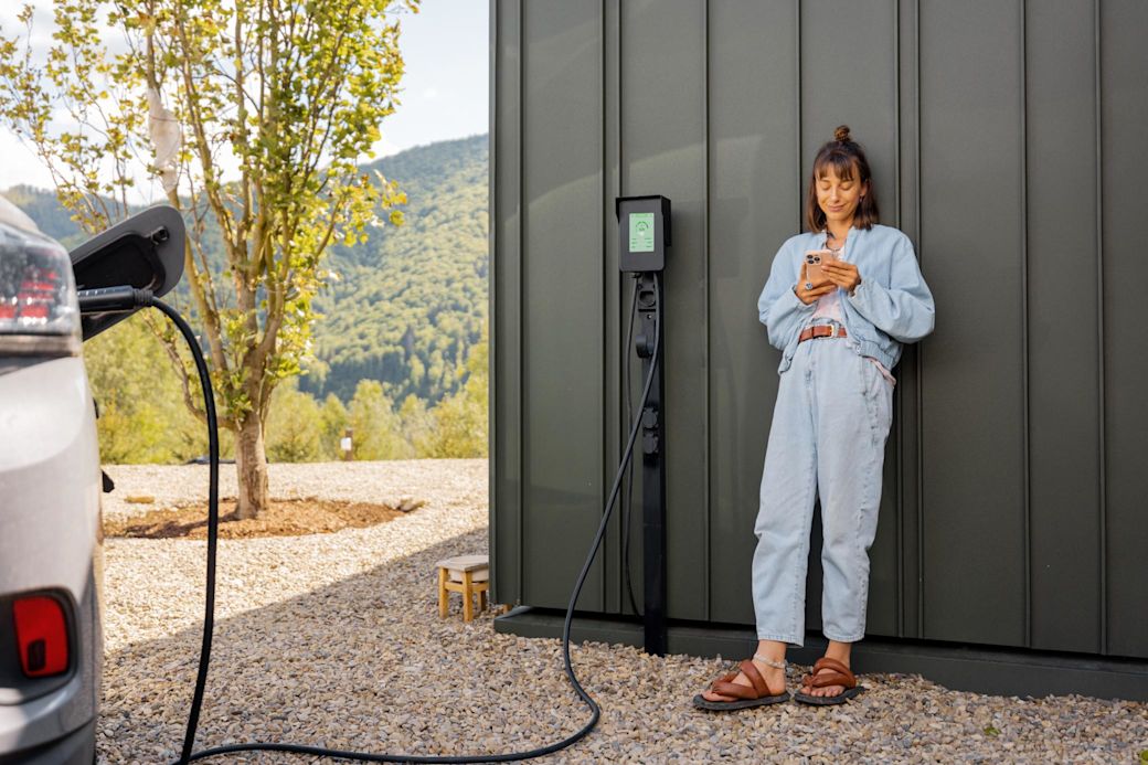 Junge Frau lehnt an Garage und hält ein Handy in der Hand während sie ihr E-Auto zuhause mit Wallbox lädt