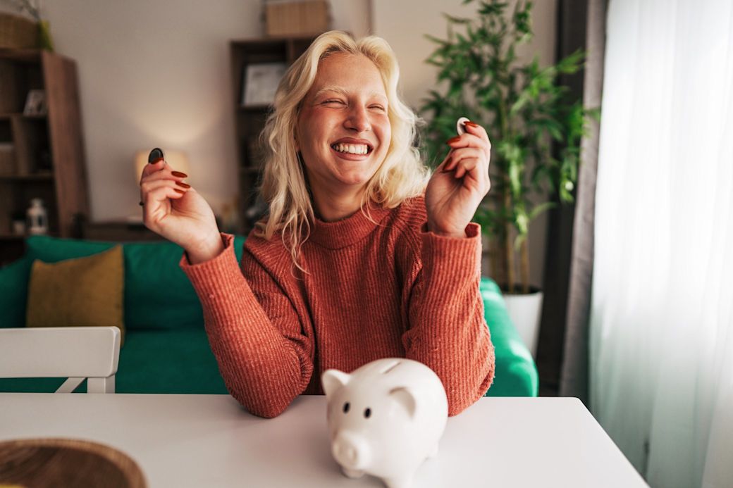 Blonde Frau mit rotem Pullover hält Münzen in der Hand und lacht vor ihrem Sparschwein auf dem Tisch.