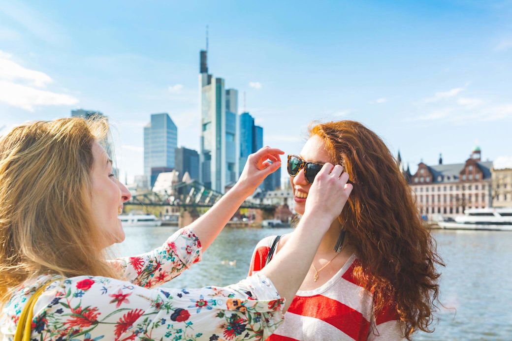 Frauen lachen zusammen im Freien, mit Sonnenbrille und spazieren auf der Uferpromenade von Frankfurt.