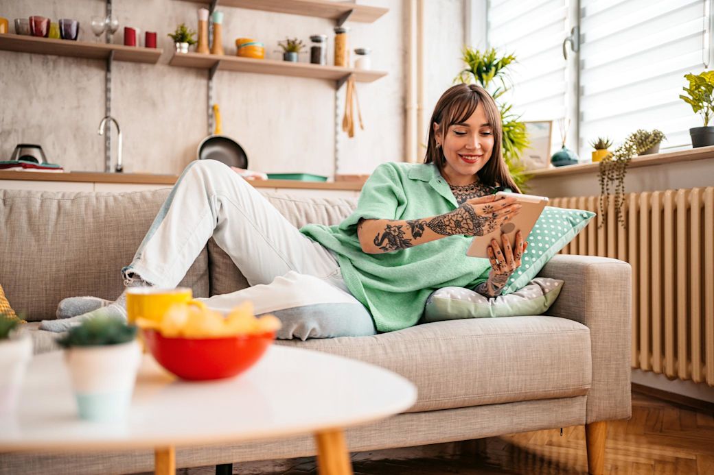 Junge Frau auf dem Sofa steuert mit dem Tablet ihre Smart-Home-Geräte