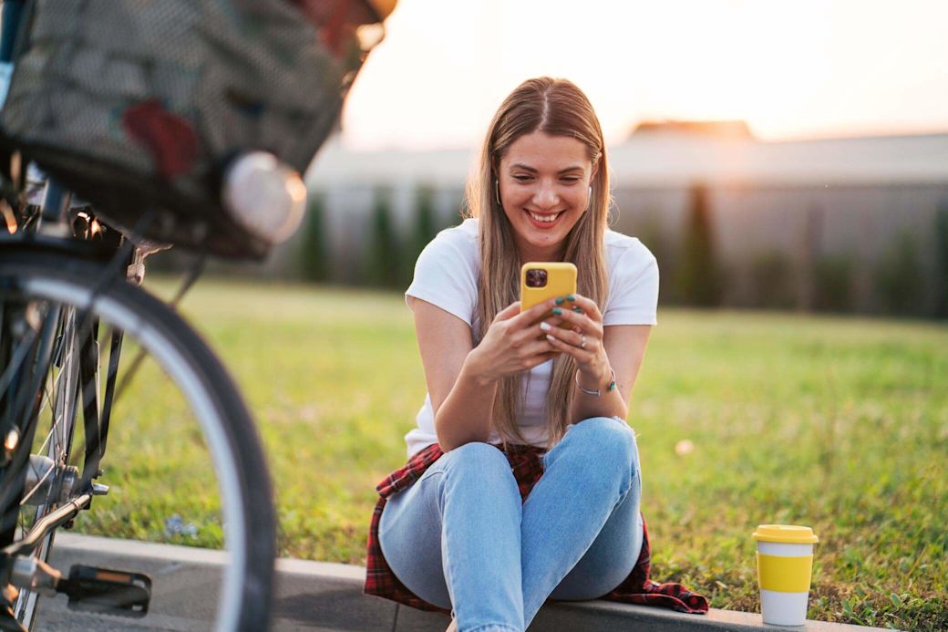 Junge Frau sitzt mit Ihrem Smartphone am Straßenrand und lächelt.