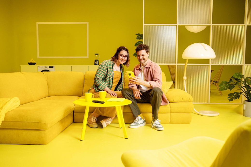 Ein Mann und eine Frau sitzen auf einem Sofa in einem gelben Wohnzimmer. Sie schauen gemeinsam auf ein Tablet und lächeln. Alles im Raum ist in Gelbtönen gehalten.