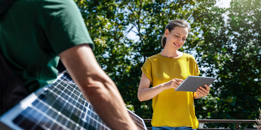 Frau im gelben Shirt steht in der Natur vor grünen Bäumen mit Tablet in der Hand und Mann mit grünem Shirt trägt ein Solarpanel