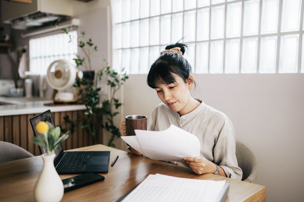 Eine junge Frau sitzt mit ihrem Laptop am Tisch und prüft Papiere und Rechnungen