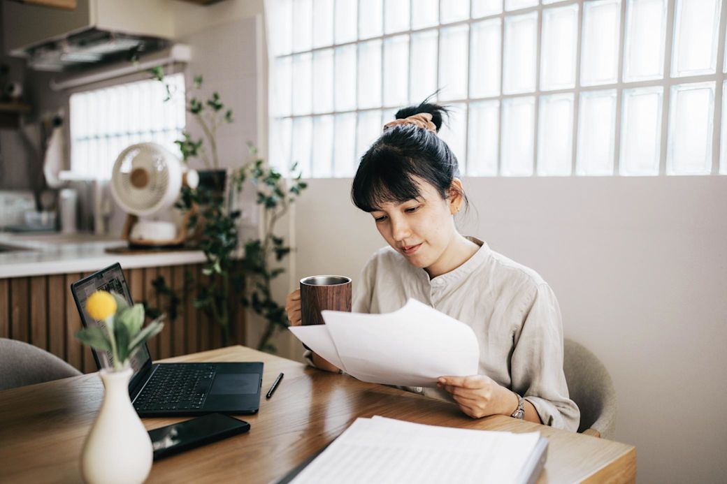 Eine junge fokussierte Frau sitzt mit ihrem Laptop an einem Tisch und liest Papiere mit Steuern, Rechnungen und Finanzen.