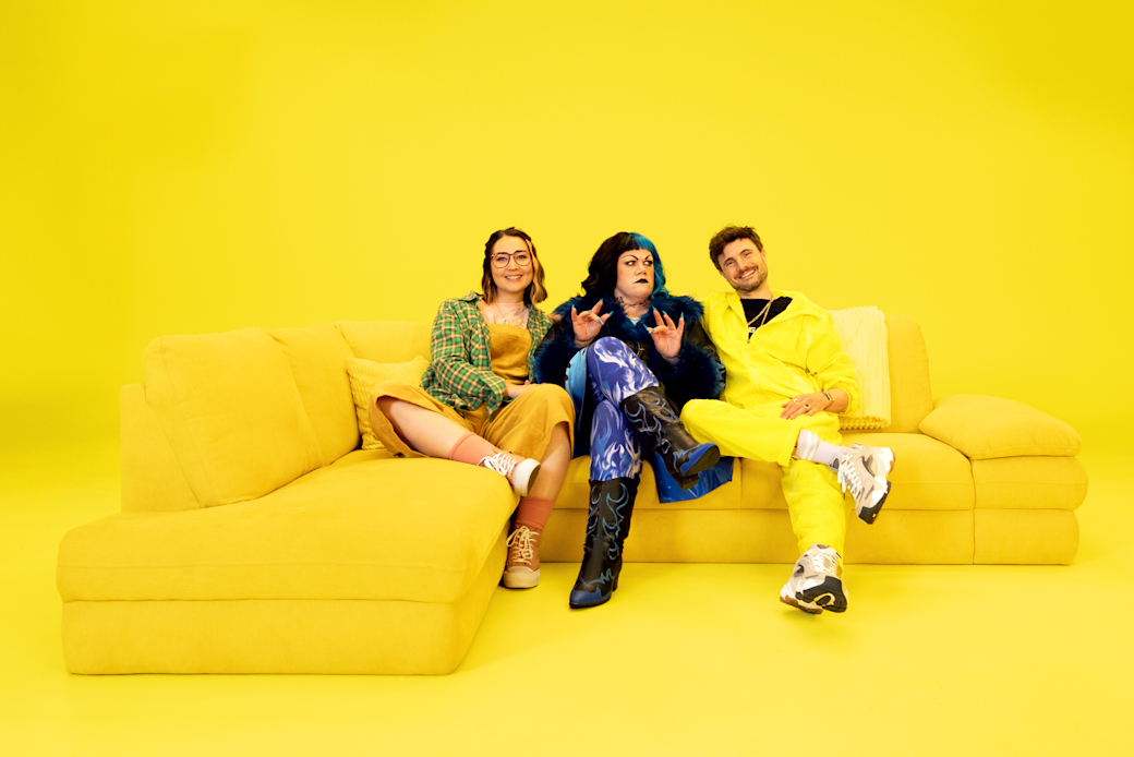 Drei Personen sitzen auf einem gelben Sofa vor gelbem Hintergrund, tragen bunte, modische Kleidung und lässige Schuhe.