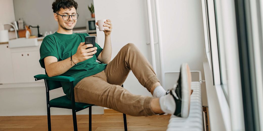 Junger Mann sitzt lächelnd am Handy, mit den Füßen auf der Heizung