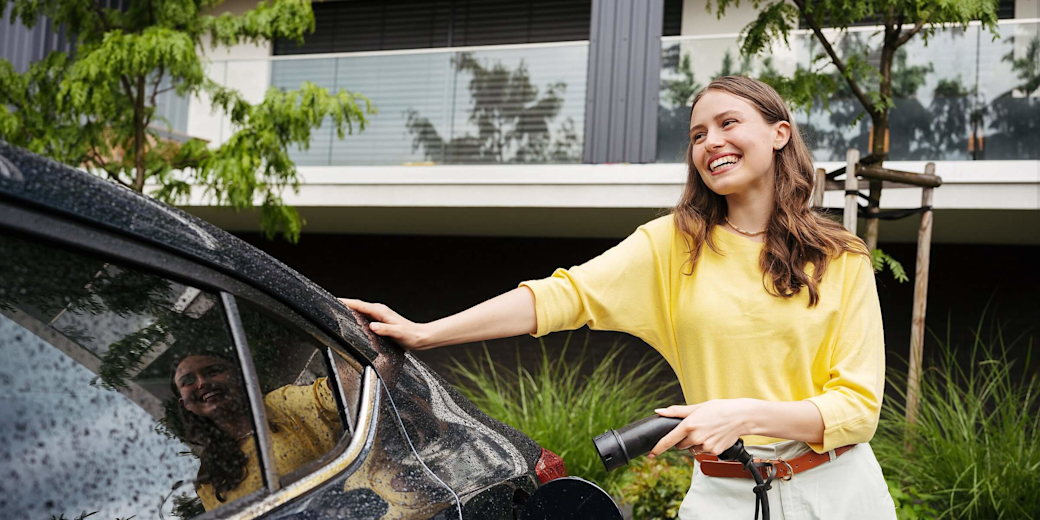 Lachende Frau im gelben Pullover lädt ihr E-Auto zuhause
