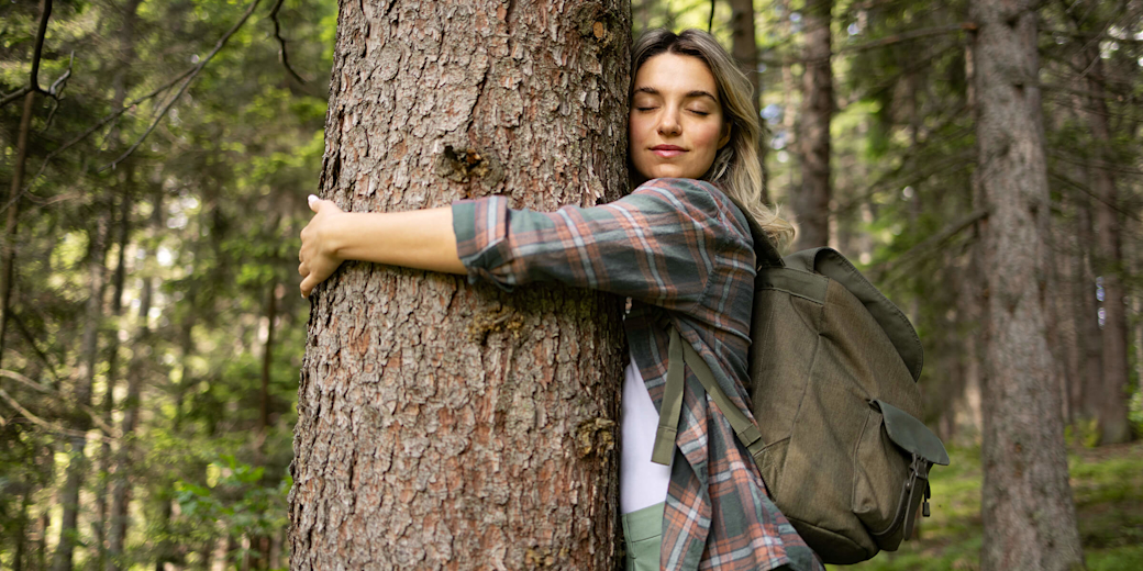 Frau steht im Wald und umarmt einen Baum.