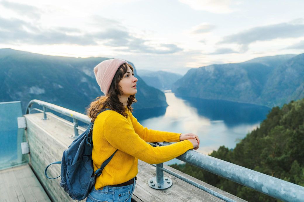 Junge Frau mit rosa Mütze und gelbem Pullover blickt auf Berge und Wasser am Wasserkraftwerk