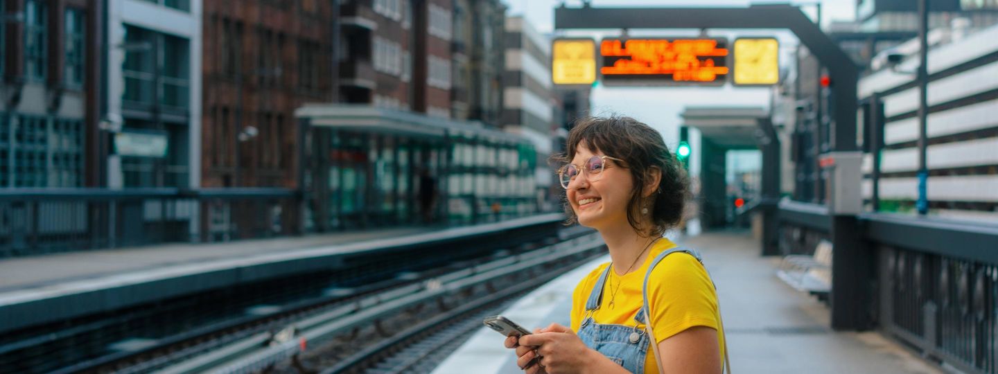 Frau in einem gelben TShirt wartet mit ihrem Handy an einem Bahnsteig