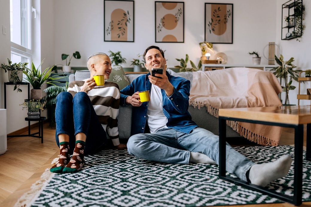 Ein junges Paar sitzt mit einer Tasse Tee in der Hand auf dem Boden und steuert ein Smart-Home-System mit dem Smartphone