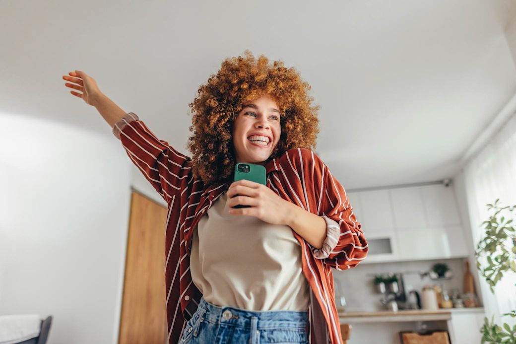 Junge Frau mit lockigem roten Haar hält ein Smartphone, während sie fröhlich in ihrer Wohnung tanzt.