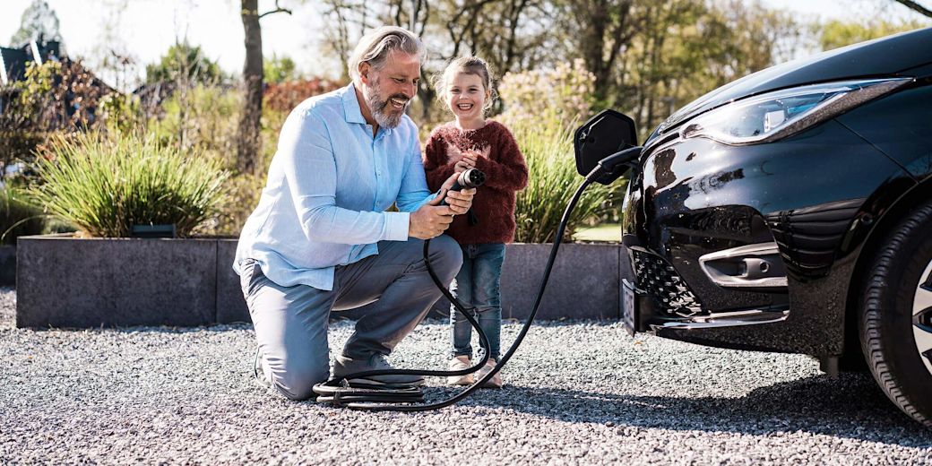 Mann mit Tochter, der an einem sonnigen Tag vor einem E-Auto einen Ladekabel in der Hand hält