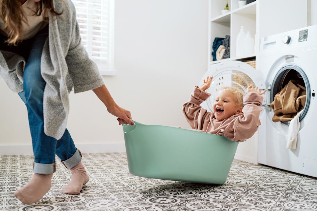 Eine Mutter vor der Waschmaschine spielt mit ihrer Tochter, die im Wäschekorb sitzt.