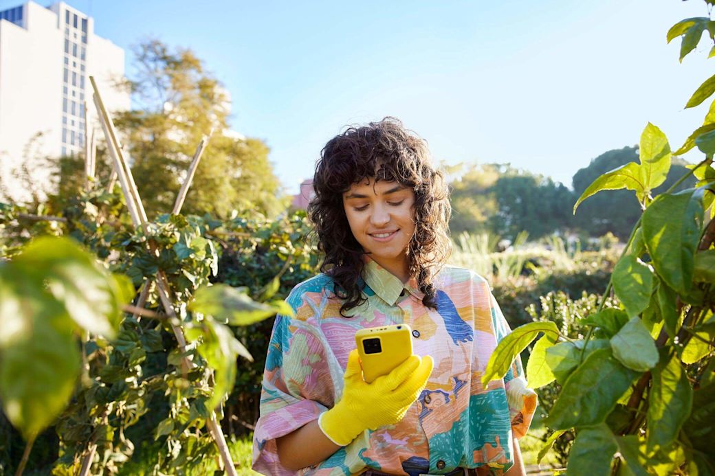 Eine Frau steht im Garten und schaut auf ihr gelbes Handy.