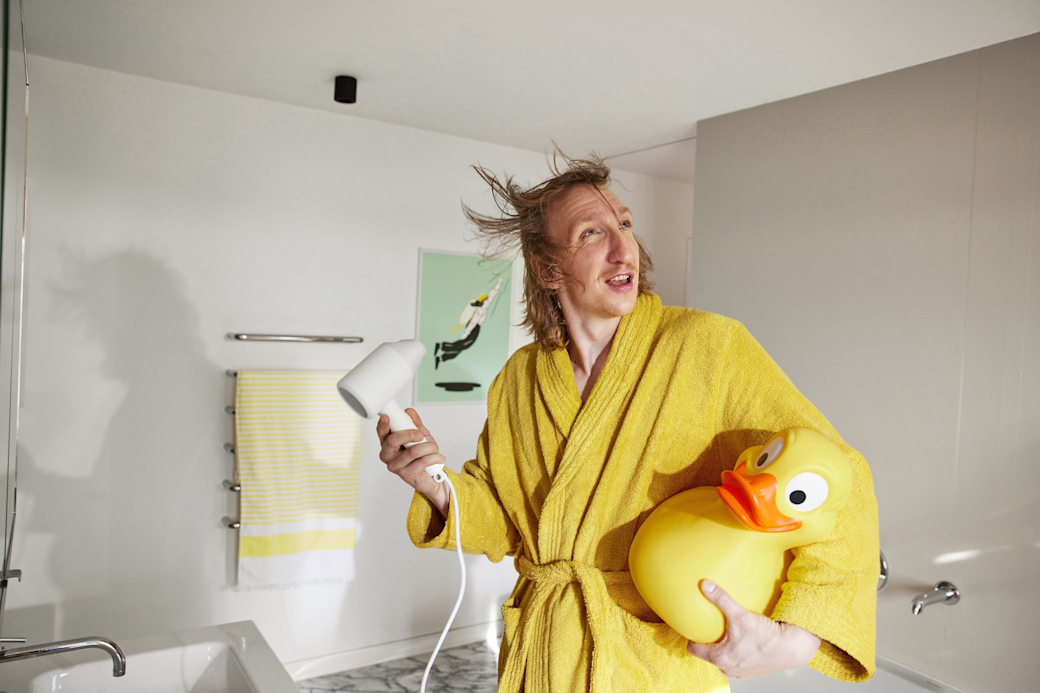 Fröhlicher Mann im Bad mit gelber Ente föhnt seine Haare