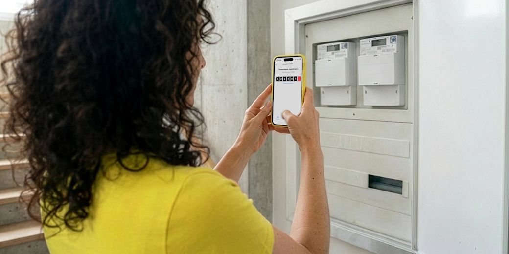 Frau mit braunen Locken und gelbem Shirt steht vor Zählerkasten und fotografiert Zählerstand mit gelbem Handy und der Yello App.