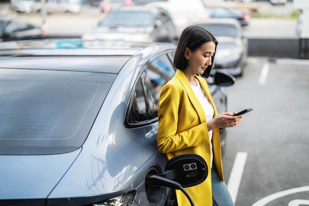 Frau in gelbem Jacket ist an ihrem Handy während das E-Auto lädt