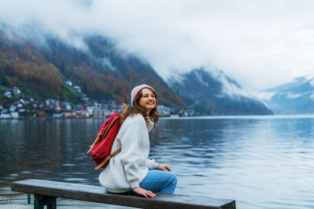 Frau sitzt mit rotem Rucksack am See und blickt in die verschneiten Berge