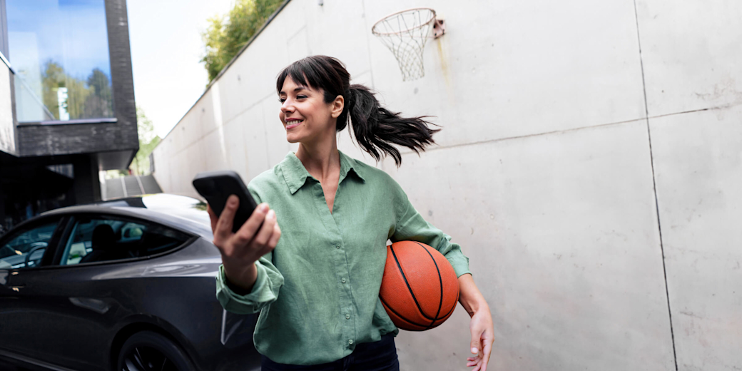 Frau mit Basketball und Handy lädt ihr E-Auto zuhause