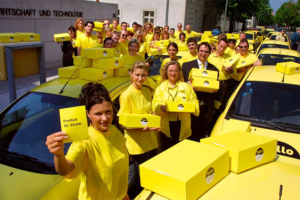 Eine Gruppe von Menschen in gelben Hemden, die gelbe Schachteln & Karten halten. Auf den Karten steht „Freiheit für Strom“. 