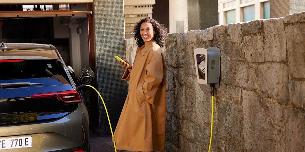 Frau lehnt an der Wand und lädt E-Auto über eine Wallbox