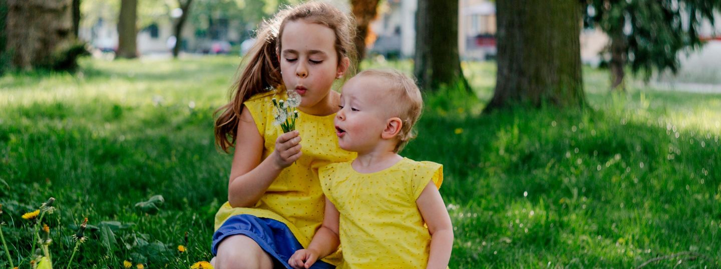 Kinder im selben gelben Outfit hocken auf einer Wiese und pusten Pusteblumen