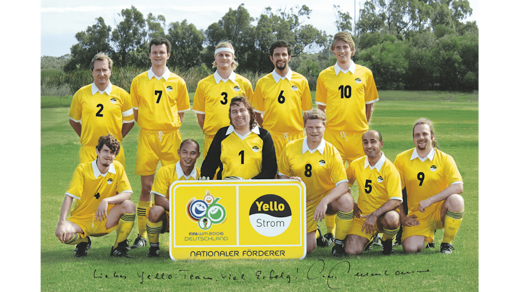 Die Yello-Fußballmannschaft in gelben Trikots posiert auf einem Feld mit einem Schild, auf dem Logos und Text zu sehen ist: „FIFA WM 2006 Deutschland“, ‚Nationaler Förderer‘ und ‚Yello Strom‘.