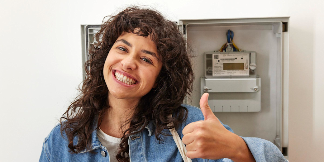 Eine Frau steht vor einem Stromzähler, lächelt, und hält ihren Daumen nach oben