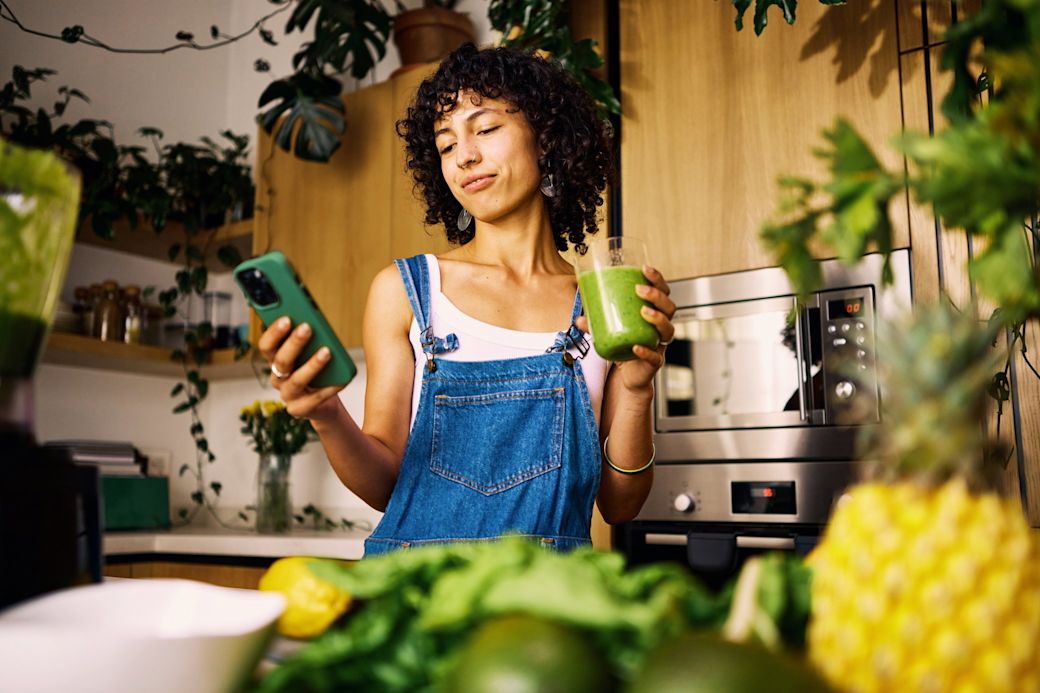Frau mit Smoothie in der Hand steht in der Küche und schaut auf ihr Smartphone